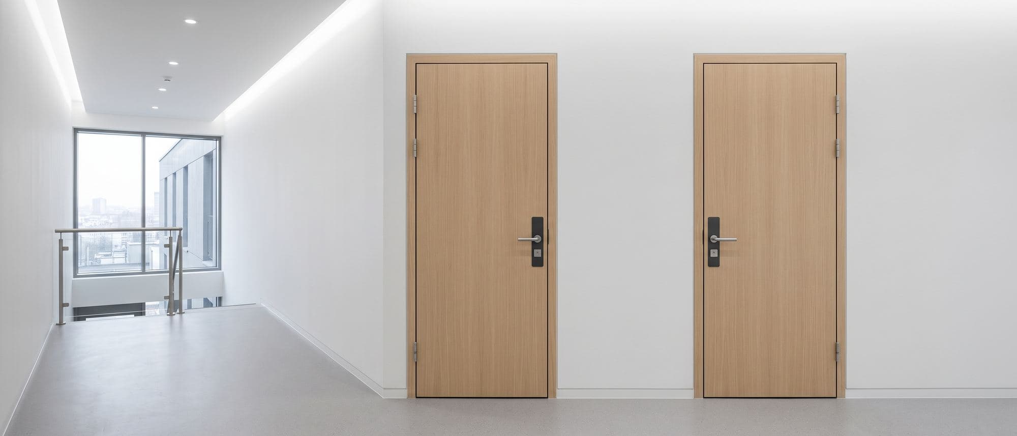 Palier d’immeuble récent, deux portes palières en chêne clair côte à côte, sol résine et éclairage encastré